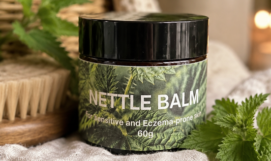 A jar of Nettle Balm with a black lid and a green leaf-patterned label, styled on a rustic wooden surface with fresh nettle leaves, a woven basket, and a soft beige blanket in a warm, blurred background.
